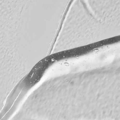 Primer plano del Solawaves LightBoost Wand Activating Serum, un gel transparente con ácido hialurónico. Presenta burbujas de aire sobre un fondo blanco texturizado, resaltando la superficie lisa y brillante en contraste con el fondo rugoso.