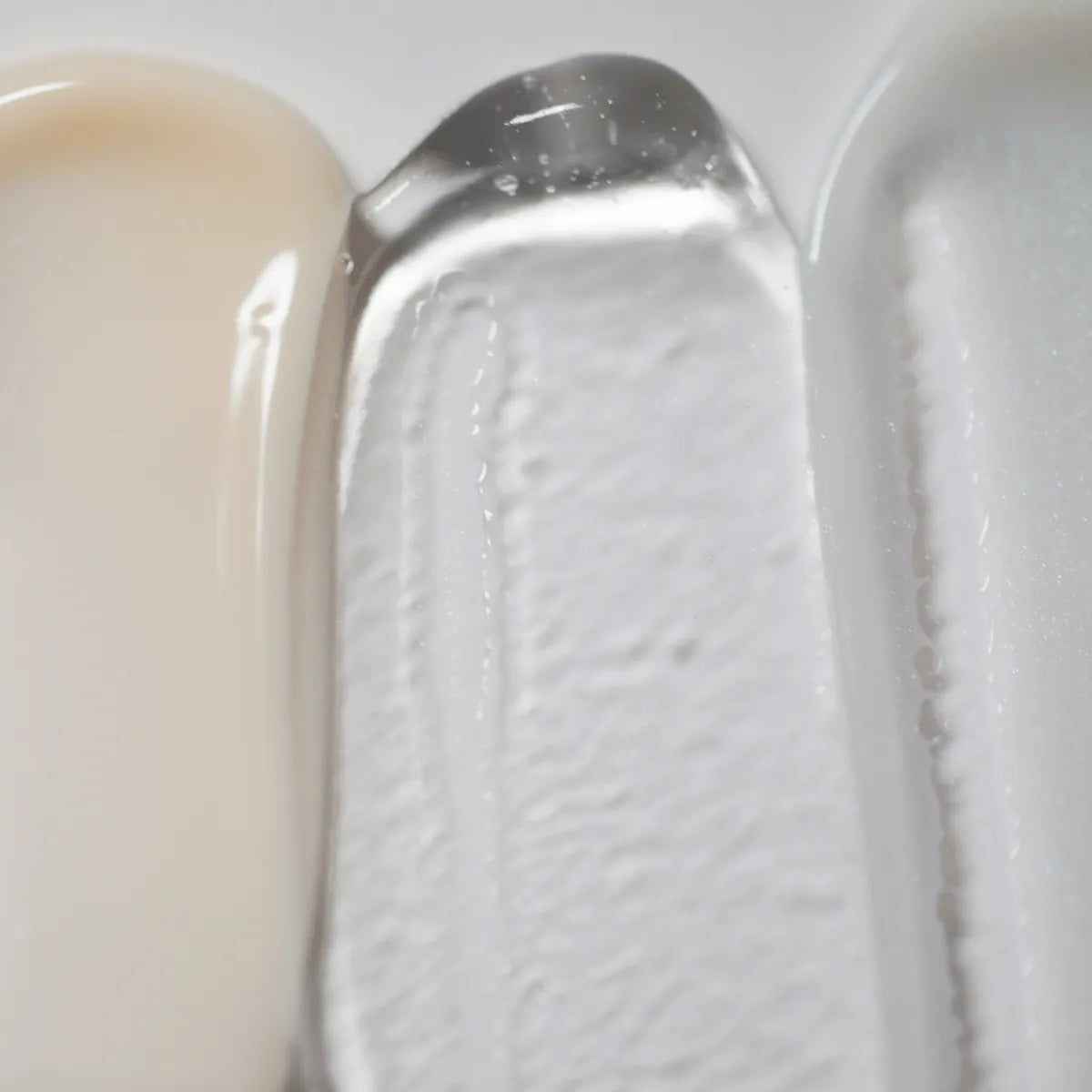 Una imagen en primer plano muestra las texturas de Solawaves LightBoost Wand Activating Serum, con beige cremoso, gel transparente con tecnología LightBoost y crema suave gris claro/blanco. Las texturas varían de cremosa a gelatinosa, cada una de ellas con un acabado brillante.